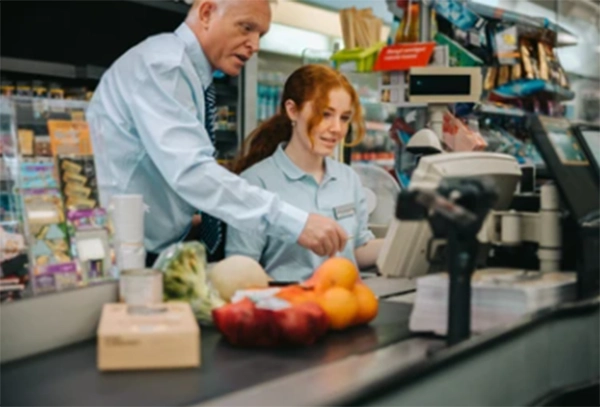 A teenage retail employee at a checkout counter while a shift manager reviews a printed schedule